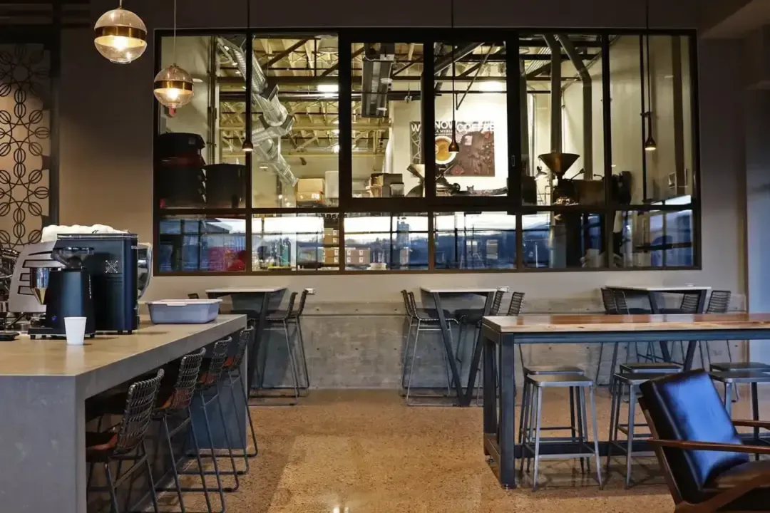 A window from the seating area looking into a large industrial room with coffee roasting equipment.