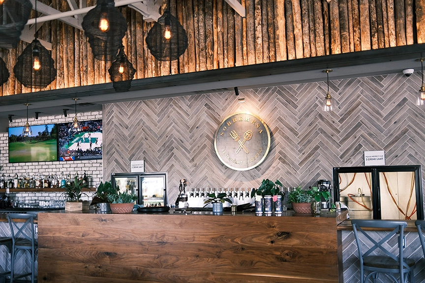 Wood panel bar facing a row of liquor bottles and beer taps, backed by warmly lit tile and tree branch lined walls.