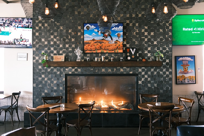 Circular wooden tables for three surrounded by dark, rustic yet simple chairs next to a large fireplace and shelf with various medals and small ornaments.